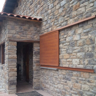 Exterior de edificio con revestimiento de pared en aplacado efecto piedra natural en tonos beige y gris, marco de madera en ventana, puerta de entrada y cornisa de barro cocido en la línea de tejado.