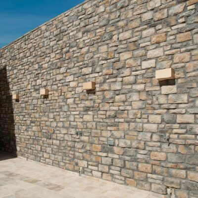 Fachada exterior revestida con aplacado de piedra natural en tonos beige y gris con textura irregular y acabado rústico. Pavimento de hormigón gris claro en base y pared complementaria de ladrillo marrón a la izquierda bajo cielo azul despejado.