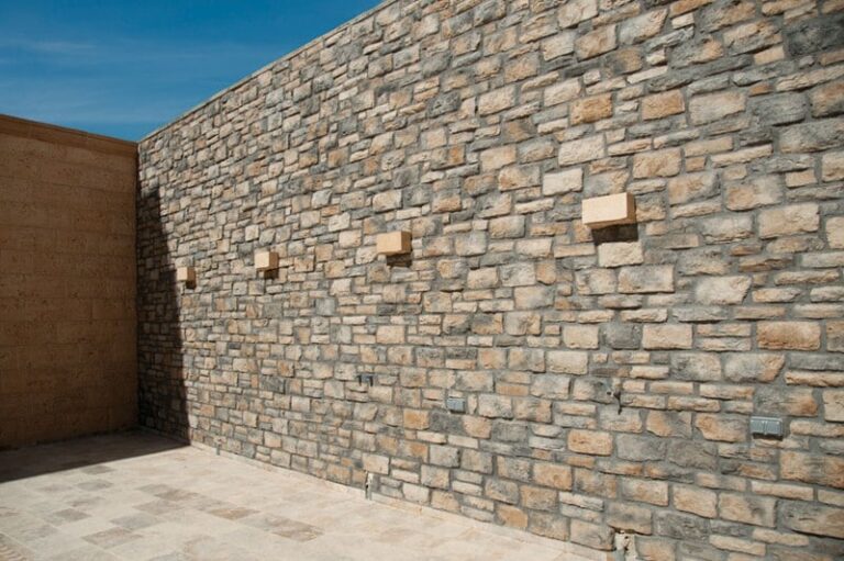 Fachada exterior revestida con aplacado de piedra natural en tonos beige y gris con textura irregular y acabado rústico. Pavimento de hormigón gris claro en base y pared complementaria de ladrillo marrón a la izquierda bajo cielo azul despejado.