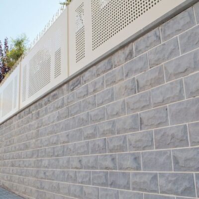 Revestimiento de pared exterior con aplacado gris efecto piedra natural en formato rectangular, instalado en fachada moderna. Superior con panel perforado beige y vegetación ornamental.