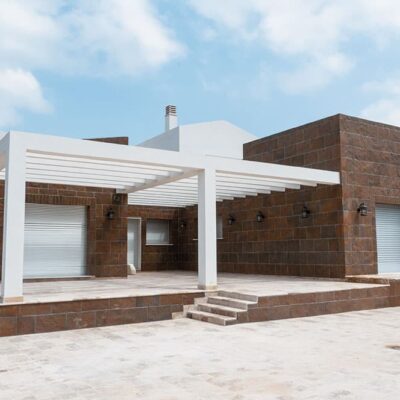 Construcción contemporánea con muros de revestimiento pizarra cobrizo en tonos marrones rojizos, acabado mate rugoso. Pórtico blanco minimalista y suelos porcelanicos de pavimento claro en la entrada elevada, con montañas visibles al fondo.