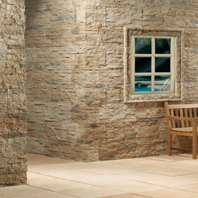 Terraza exterior con revestimiento de piedra natural Plasencia Castaño en tonos beige y arena. Suelo porcelánico imitación piedra, banco de madera, columna cilíndrica y ventana con marco de piedra.