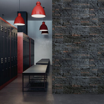 Pasillo con revestimiento de pared tipo piedra gris oscura en formato modular, suelo de cemento pulido, bancos negros minimalistas y tres lámparas colgantes rojas industrial. El revestimiento presenta textura rústica característica del gres porcelanico.