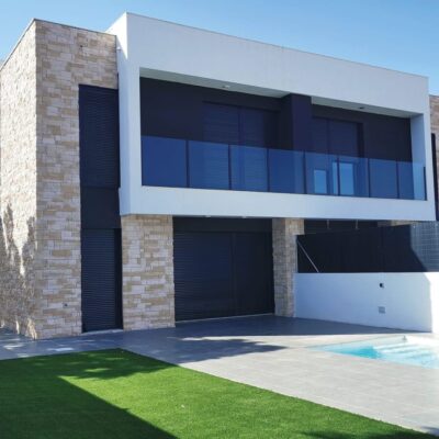 Vivienda contemporánea de dos plantas con revestimiento de fachada en tono piedra natural gris claro, combinado con paneles blancos y grandes ventanales azules. El aplacado efecto piedra cubre las columnas verticales del edificio, creando un contraste elegante con la arquitectura minimalista.