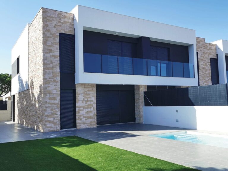 Vivienda contemporánea de dos plantas con revestimiento de fachada en tono piedra natural gris claro, combinado con paneles blancos y grandes ventanales azules. El aplacado efecto piedra cubre las columnas verticales del edificio, creando un contraste elegante con la arquitectura minimalista.