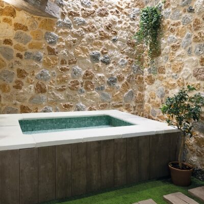 Piscina rectangular con revestimiento de azulejos verde agua y borde blanco, rodeada de paredes de piedra natural beige y plantas verdes en maceta. El diseño combina acabados rústicos con líneas modernas.