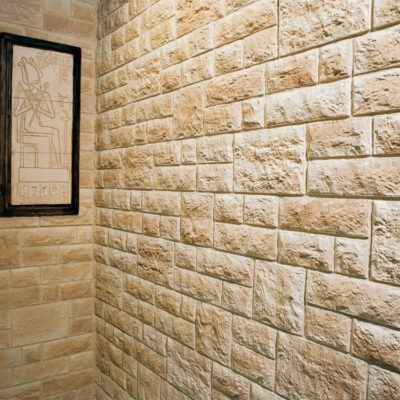 Rincón de baño con paredes revestidas en travertino split con acabado natural texturizado en tonos beige y crema. Un marco con grabado egipcio decora la pared lateral en contraste con el aplacado de piedra.