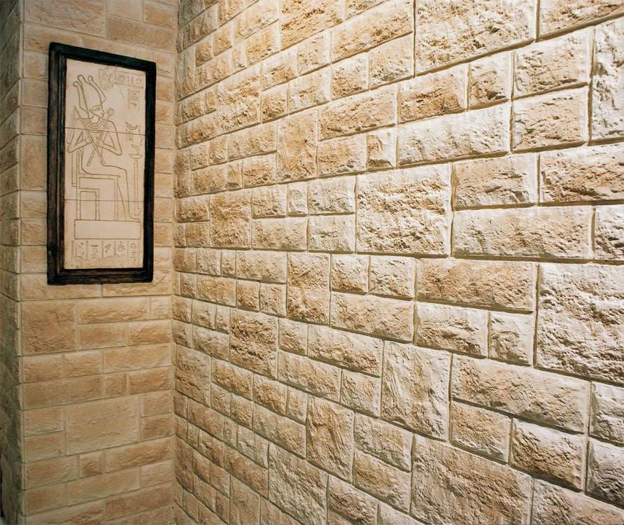 Rincón de baño con paredes revestidas en travertino split con acabado natural texturizado en tonos beige y crema. Un marco con grabado egipcio decora la pared lateral en contraste con el aplacado de piedra.