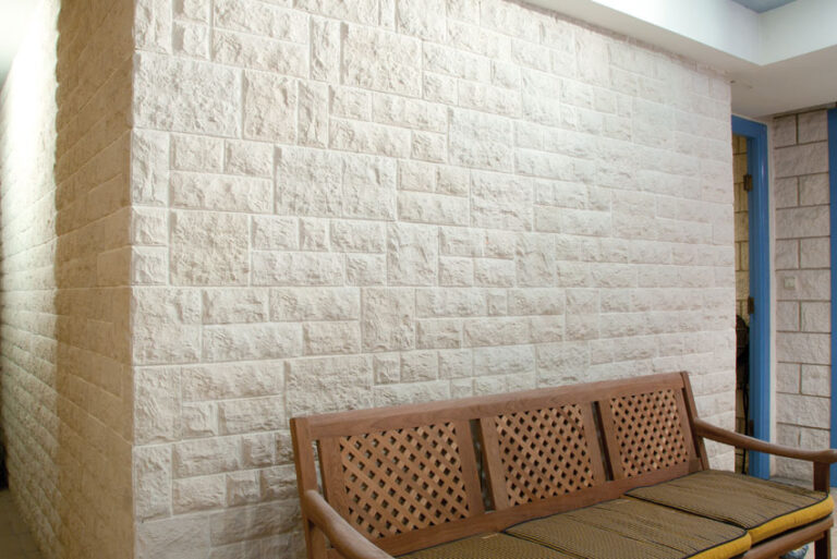 Pared de salón revestida con baldosas de hormigón efecto travertino en tonos crudo y beige, con textura split rugosa. Sofá de madera con respaldo perforado y asiento tapizado en primer plano.