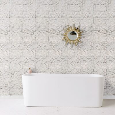 Baño contemporáneo con bañera blanca frente a pared revestida con azulejos de textura clara y tonos naturales. Espejo circular dorado con acabado sunburst colgado sobre el revestimiento.