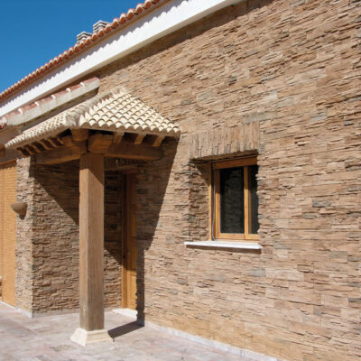 Fachada exterior de vivienda con revestimiento de pared imitación piedra en tonos cálidos y beige, puerta de entrada con dintel de madera y ventana con marco de madera, bajo un cielo despejado.