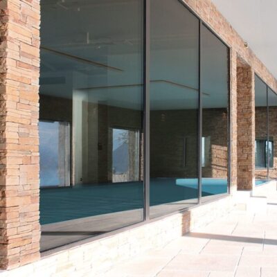 Terraza exterior con suelo porcelánico de gran formato en tonos claros, columnas de ladrillo visto, amplios ventanales de vidrio y piscina azul. El revestimiento de suelo ceramico cubre la zona de descanso con acabado antideslizante.