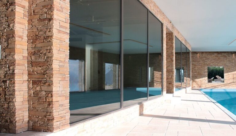 Terraza exterior con suelo porcelánico de gran formato en tonos claros, columnas de ladrillo visto, amplios ventanales de vidrio y piscina azul. El revestimiento de suelo ceramico cubre la zona de descanso con acabado antideslizante.