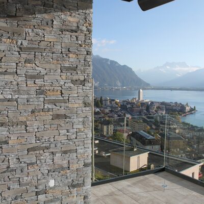 Terraza exterior con aplacado de piedra gris en pared lateral, piso de hormigón y barandilla de cristal. Al fondo, vistas panorámicas de un lago alpino rodeado de montañas y un pueblo ribereño bajo cielo despejado.