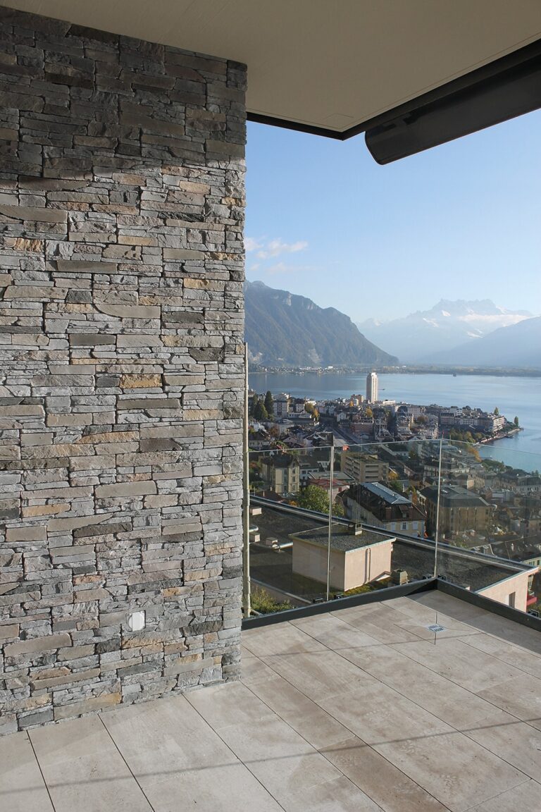 Terraza exterior con aplacado de piedra gris en pared lateral, piso de hormigón y barandilla de cristal. Al fondo, vistas panorámicas de un lago alpino rodeado de montañas y un pueblo ribereño bajo cielo despejado.