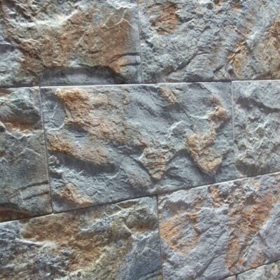 Superficie de revestimiento con textura rústica de piedra en tonos grises y beige, acabado mate con relieve irregular y veteado natural que simula mampostería de piedra natural.
