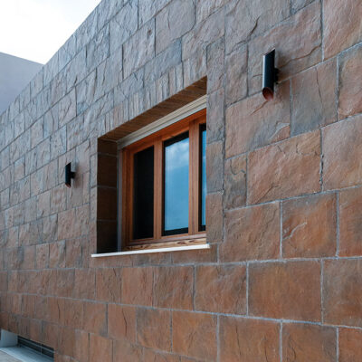Fachada exterior construida con revestimiento de pizarra cobrizo en tonos tostados y grises. Los bloques de piedra natural se distribuyen en formato rectangular, mostrando la textura rugosa característica y variaciones de color que conforman un acabado mate en toda la superficie.