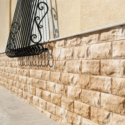 Fachada exterior con revestimiento de imitación piedra en tonos beige y crema, instalado en formato rectangular. La pared muestra un acabado rustico con textura de piedra natural, complementado con una moldura de acero decorativa en la parte superior.