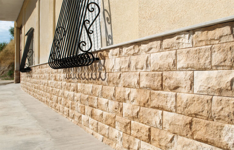 Fachada exterior con revestimiento de imitación piedra en tonos beige y crema, instalado en formato rectangular. La pared muestra un acabado rustico con textura de piedra natural, complementado con una moldura de acero decorativa en la parte superior.