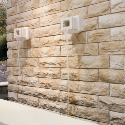Esquina exterior de edificio con revestimiento de pared Saboya Aneto en tonos beige y crema, mostrando bloques rectangulares de piedra natural con textura rústica mate. Accesorios blancos montados en la superficie y jardín de fondo.