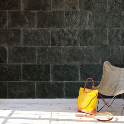 Espacio interior luminoso con pared de revestimiento de pizarra en tonos gris oscuro y formato rectangular. Una silla butterfly beige y accesorios de ocio completan la escena junto a una ventana que proyecta luz natural.