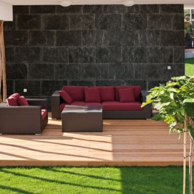 Terraza exterior con pared de revestimiento de pizarra gris oscuro en formato rectangular, equipada con mobiliario de jardín de ratán marrón y cojines rojo burdeos. La zona está pavimentada con tarima de madera clara y conecta con el jardín de césped verde.