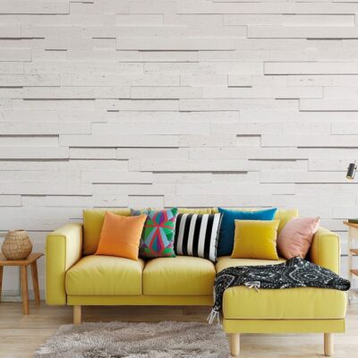 Salón contemporáneo con pared de revestimiento decorativo imitación piedra blanca Tivoli en acabado crudo. Sofá amarillo con cojines multicolores y ottoman a juego sobre suelo de madera clara.