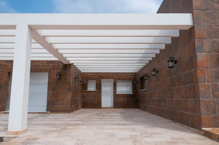 Terraza cubierta con estructura de vigas blancas y paredes revestidas en pizarra cobrizo de gran formato. El suelo cerámico gris complementa el acabado rústico de la cladding de piedra natural.