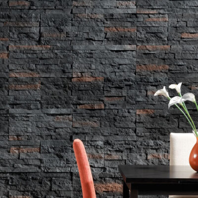 Sala de estar con pared de revestimiento Plasencia Vulcano en tonos gris oscuro y marrón rojizo con acabado de piedra volcánica apilada. Una silla de tela coral y una mesa negra con flores blancas en jarrón complementan el espacio.