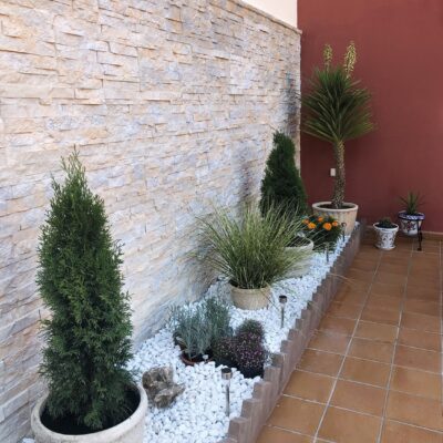 Patio exterior con revestimiento de piedra natural en tonos castaño claro instalado en pared, decorado con plantas ornamentales en macetas de hormigón, grava blanca y suelo de gres en tonalidad terracota.
