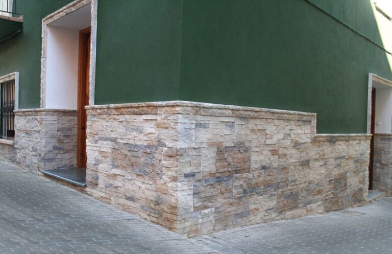 Fachada exterior con revestimiento de piedra natural en tonos castaño y gris, instalado sobre base de mortero, combinado con pared verde y puerta de madera. Ejemplo de aplicación de porcelánico imitación piedra en envolvente de edificio.