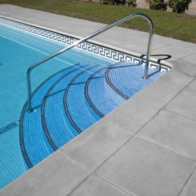 Área de piscina exterior con escaleras de entrada y barandilla metálica. El suelo alrededor de la piscina está pavimentado con grandes losas de gres porcelánico gris claro, mientras que el interior de la piscina presenta un revestimiento de mosaico azul con motivos geométricos. Al fondo se ve césped bien mantenido.