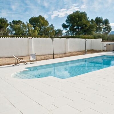 Piscina rectangular con agua turquesa rodeada de pavimento porcelánico en tonos blancos y grises claros. Muro de hormigón blanco con remate decorativo, tumbonas de madera, y zona ajardinada con árboles al fondo bajo cielo despejado.