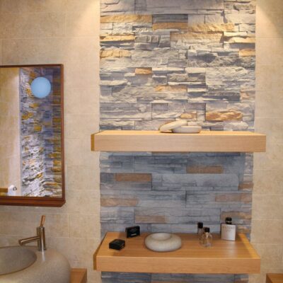 Baño contemporáneo con aplacado de piedra interior gris en tonos grises y dorados formando un muro de revestimiento texturizado detrás de dos encimeras de madera natural con lavabos redondos de cerámica.