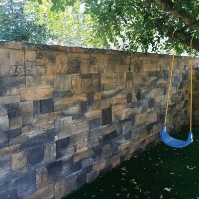 Aplacado de piedra para exterior instalado en muro de jardín, con piezas de formato irregular en tonos tierra, gris y marrón que crean un patrón de mampostería rústica. Acabado natural con textura de piedra, bajo cobertura vegetal y junto a columpio azul.