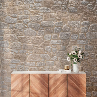 Muro de aplacado imitación piedra cortijo natur con acabado natural beige y gris, complementado con un mueble de madera clara y flores blancas decorativas sobre su superficie.