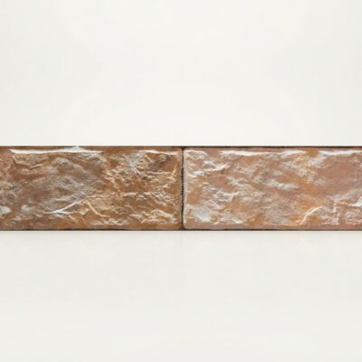 Dos piezas rectangulares de azulejo con acabado texturizado que imita piedra natural en tonos beige y grisáceos. Superficie rugosa mate con variaciones de color y efecto envejecido.