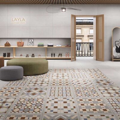 Interior de tienda concept Layla con suelo porcelánico imitación piedra en tonos beige, gris y marrón con patrones geométricos variados. Estanterías de madera con bolsos, zapatos y complementos, puffs redondos verde oliva y gris, puerta de madera con cristal hacia fachada, y plantas decorativas.