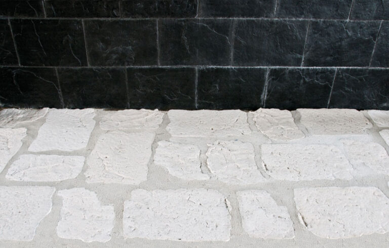 Baldosas rectangulares de gres antideslizante con acabado rústico en tonos beige y gris claro, superficie texturizada que imita piedra natural, formato suelo cerámico.
