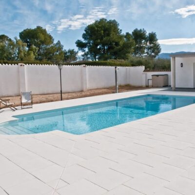 Piscina rectangular con agua turquesa rodeada de suelo porcelánico antideslizante en tonos blancos y grises claros. Muro perimetral blanco, zona de descanso con hamacas y casetas auxiliares, árboles al fondo y cielo despejado.