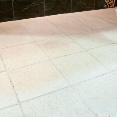 Suelo interior de baldosa de hormigón en tonos beige y crema con acabado mate y textura de piedra natural. Las piezas rectangulares están colocadas en formato métrico, mostrando la pared de fondo oscura y complementos decorativos.