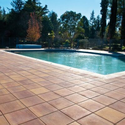 Terraza exterior con baldosa de gres en tonos cálidos junto a una piscina de agua turquesa, rodeada de árboles y vegetación en un ambiente de zona de descanso.