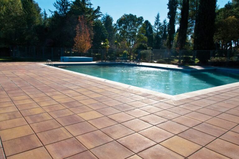 Terraza exterior con baldosa de gres en tonos cálidos junto a una piscina de agua turquesa, rodeada de árboles y vegetación en un ambiente de zona de descanso.