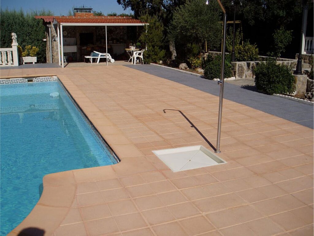 Terraza junto a piscina con suelo de baldosa terracota en tonos cálidos, mostrando el acabado rústico del material. Al fondo, caseta de piscina y zona ajardinada con vegetación.