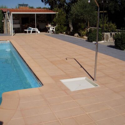 Terraza junto a piscina con suelo de baldosa terracota en tonos cálidos, mostrando el acabado rústico del material. Al fondo, caseta de piscina y zona ajardinada con vegetación.