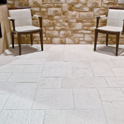 Espacio exterior pavimentado con baldosa Salinas Solaneta en tonos beige y crema, con acabado texturizado efecto piedra natural. Dos sillas de madera con tapicería blanca se sitúan sobre el suelo, frente a un muro de piedra sin desbastar y elementos arquitectónicos de piedra caliza.