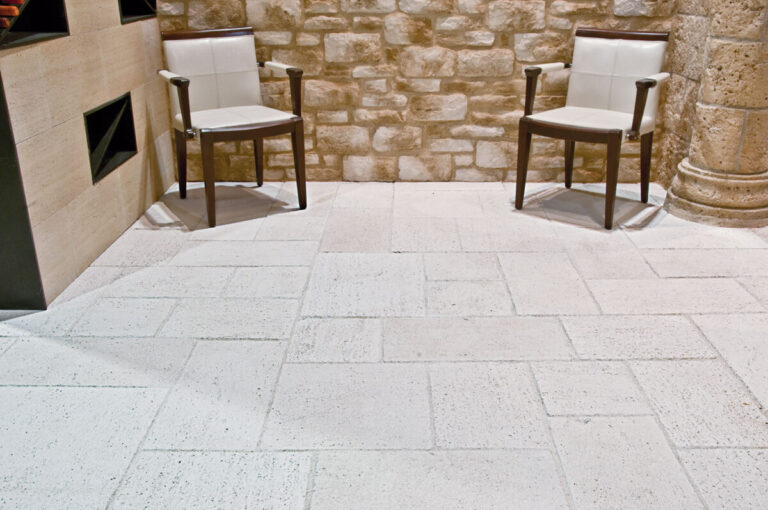 Espacio exterior pavimentado con baldosa Salinas Solaneta en tonos beige y crema, con acabado texturizado efecto piedra natural. Dos sillas de madera con tapicería blanca se sitúan sobre el suelo, frente a un muro de piedra sin desbastar y elementos arquitectónicos de piedra caliza.