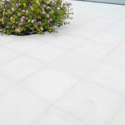 Terraza exterior pavimentada con baldosas porcelánicas de gran formato en blanco, acabado liso mate. Macetero con flores violetas y verdes como elemento decorativo sobre el suelo cerámico.