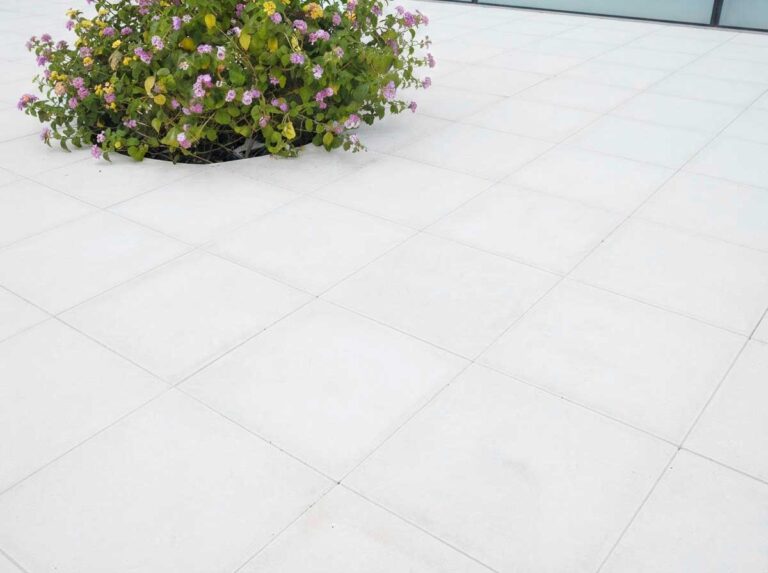 Terraza exterior pavimentada con baldosas porcelánicas de gran formato en blanco, acabado liso mate. Macetero con flores violetas y verdes como elemento decorativo sobre el suelo cerámico.
