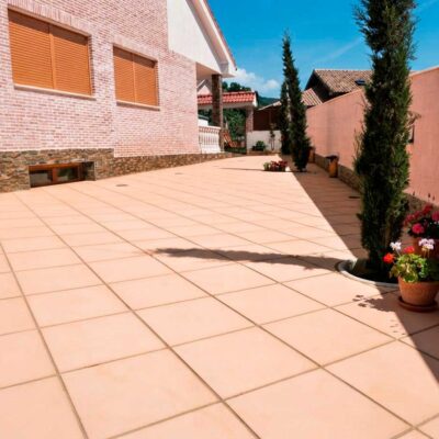 Terraza exterior pavimentada con baldosa porcelánica de gran formato en tonos beige claro, con acabado liso. La superficie muestra un patrón de rejuntado regular. Flanquean el espacio macetas de barro con flores rojas y cipreses, y se observan construcciones de ladrillo y hormigón al fondo bajo cielo despejado.